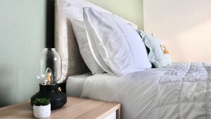 Un lit bien fait, avec une literie et des oreillers blancs, une table de chevet avec une lampe et une petite plante, et un mur vert en arrière-plan.