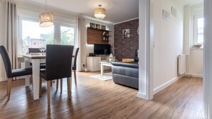 A modern living room with a dining area, a grey sofa, a TV, and wooden flooring. - Ferienwohnung in Westerdeichstrich (Westerdeichstrich)