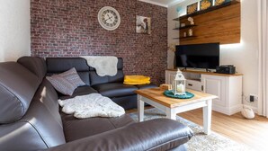 A living room with a brick wall, a black leather sofa, a wooden coffee table, and a flat-screen TV on a wooden stand. - Ferienwohnung in Westerdeichstrich (Westerdeichstrich)