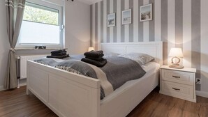 A neatly arranged bedroom with a bed, bedside table, and a window with curtains. - Ferienwohnung in Westerdeichstrich (Westerdeichstrich)