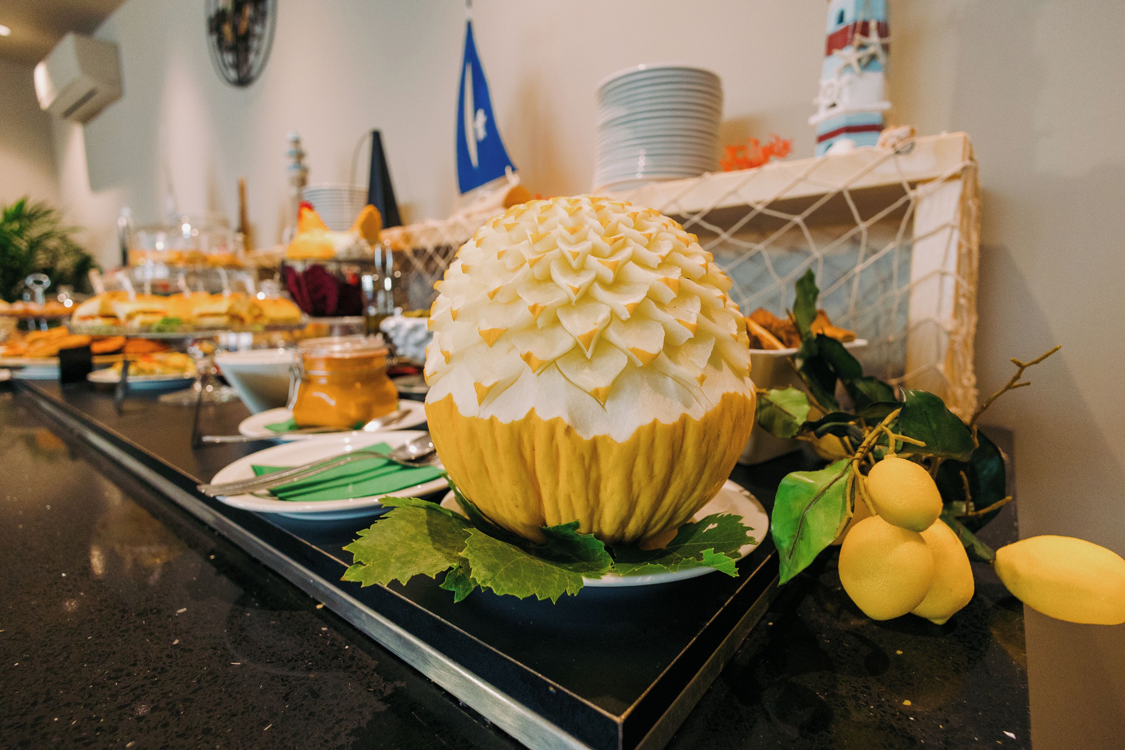 A buffet table with a large decorative fruit centerpiece, plates, and various food items.
