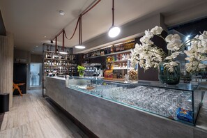 A bar area with a glass display case filled with wine glasses, a variety of bottles on shelves, and a vase with white flowers. - Baitan Hotel (Marsala)