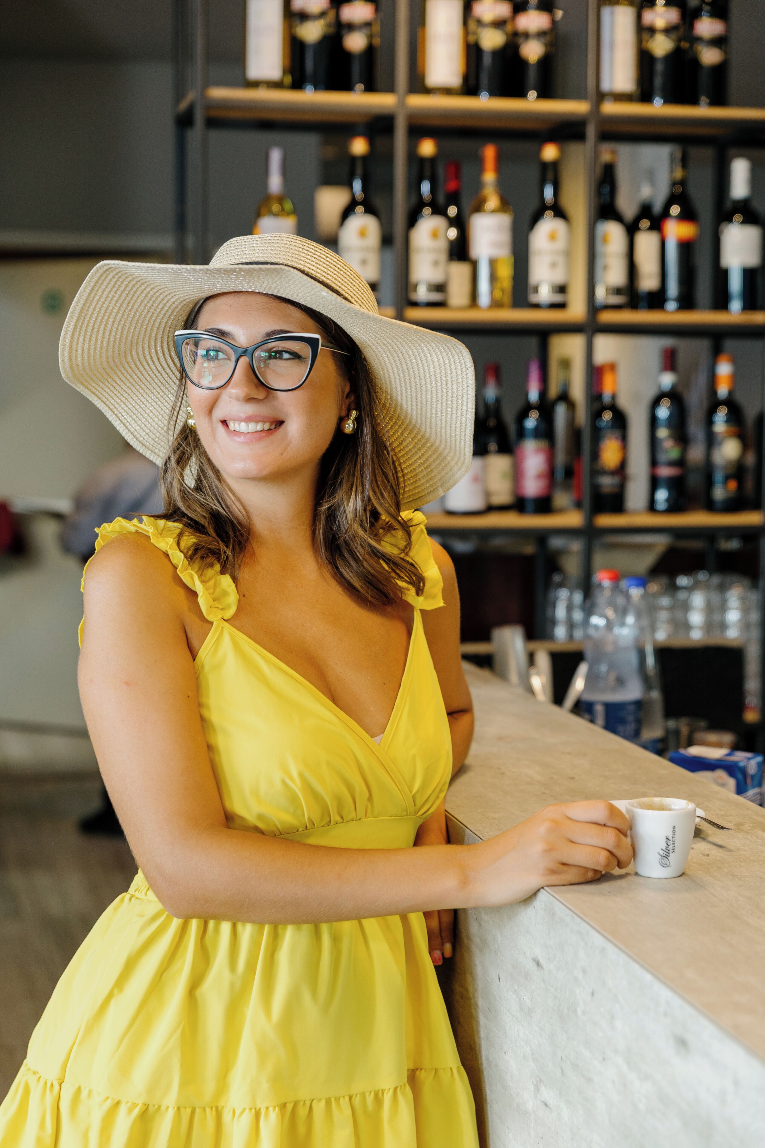 A person in a yellow dress and wide-brimmed hat leaning on a counter with a beverage cup.
