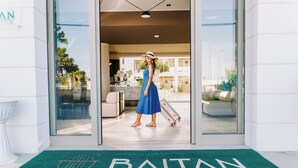 A woman in a blue dress and white hat entering a hotel with a green doormat reading "BAITAN". - Baitan Hotel (Marsala)