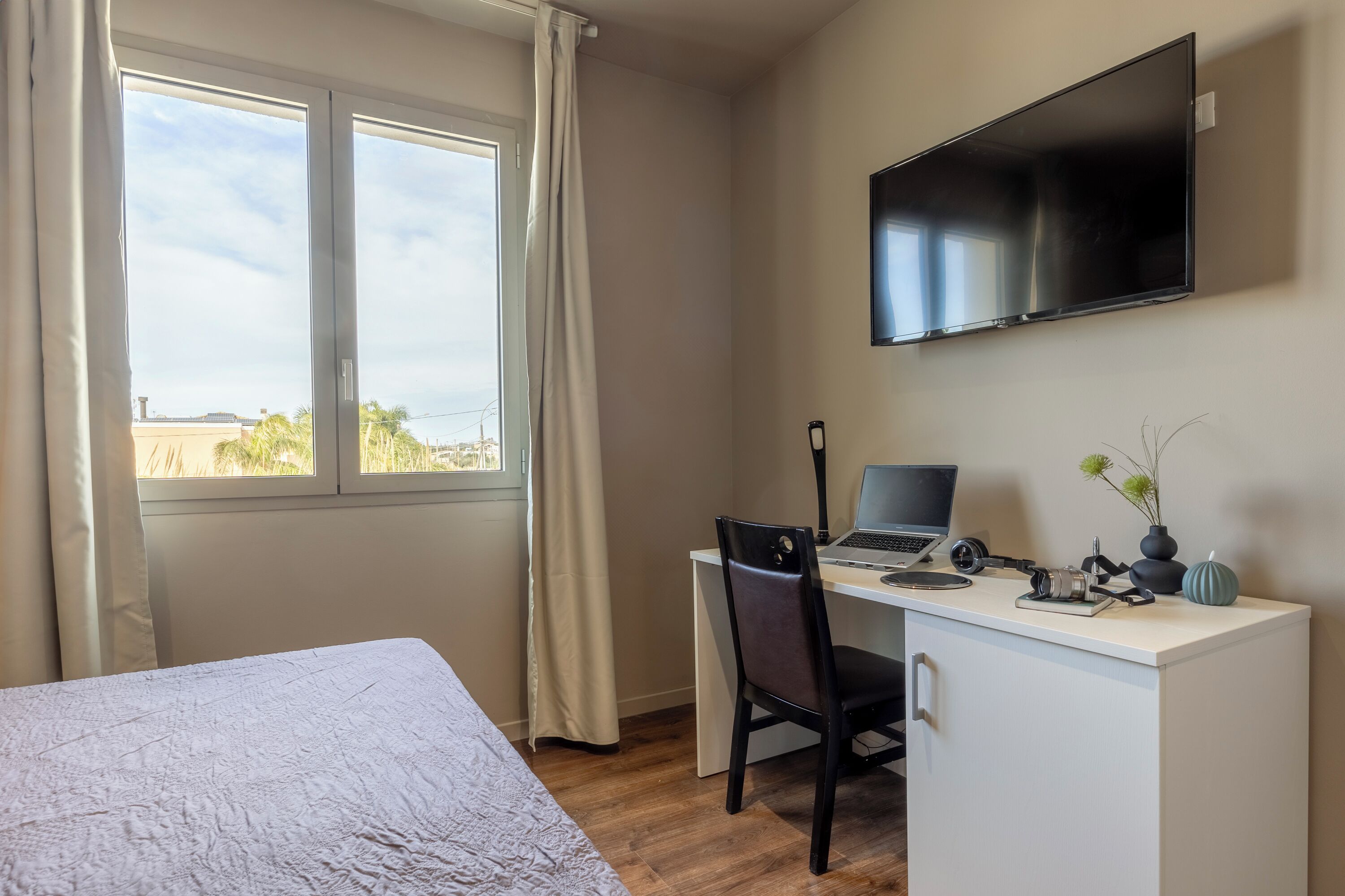 Una habitación de hotel con una cama, un escritorio con una computadora portátil, una silla, un televisor montado en la pared y una ventana con cortinas.