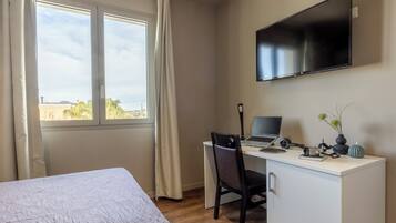 Una habitación de hotel con una cama, un escritorio con una computadora portátil, una silla, un televisor montado en la pared y una ventana con cortinas.