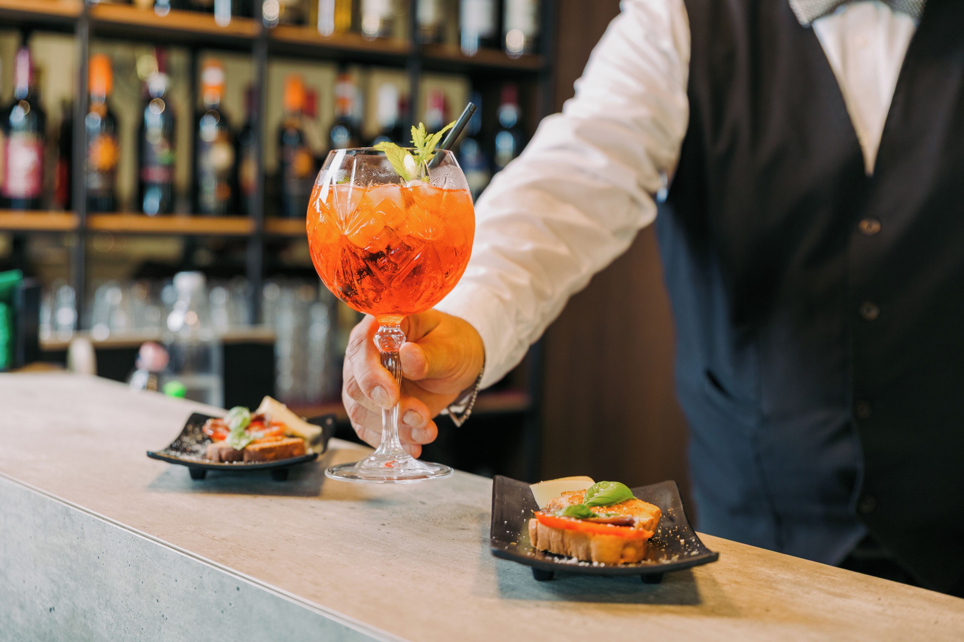 A bar setting with a bartender holding a cocktail glass, a small plate with a sandwich, and a shelf with bottles in the background.