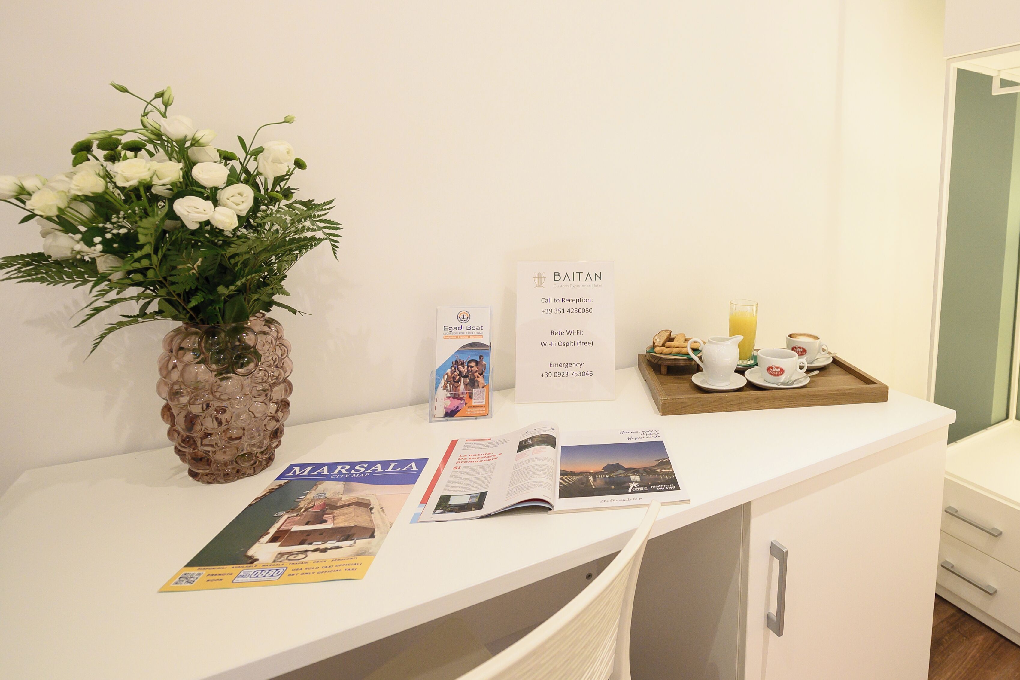 A reception desk with a vase of white flowers, informational brochures, a sign with contact details, and a tray with cups and a glass of orange juice.