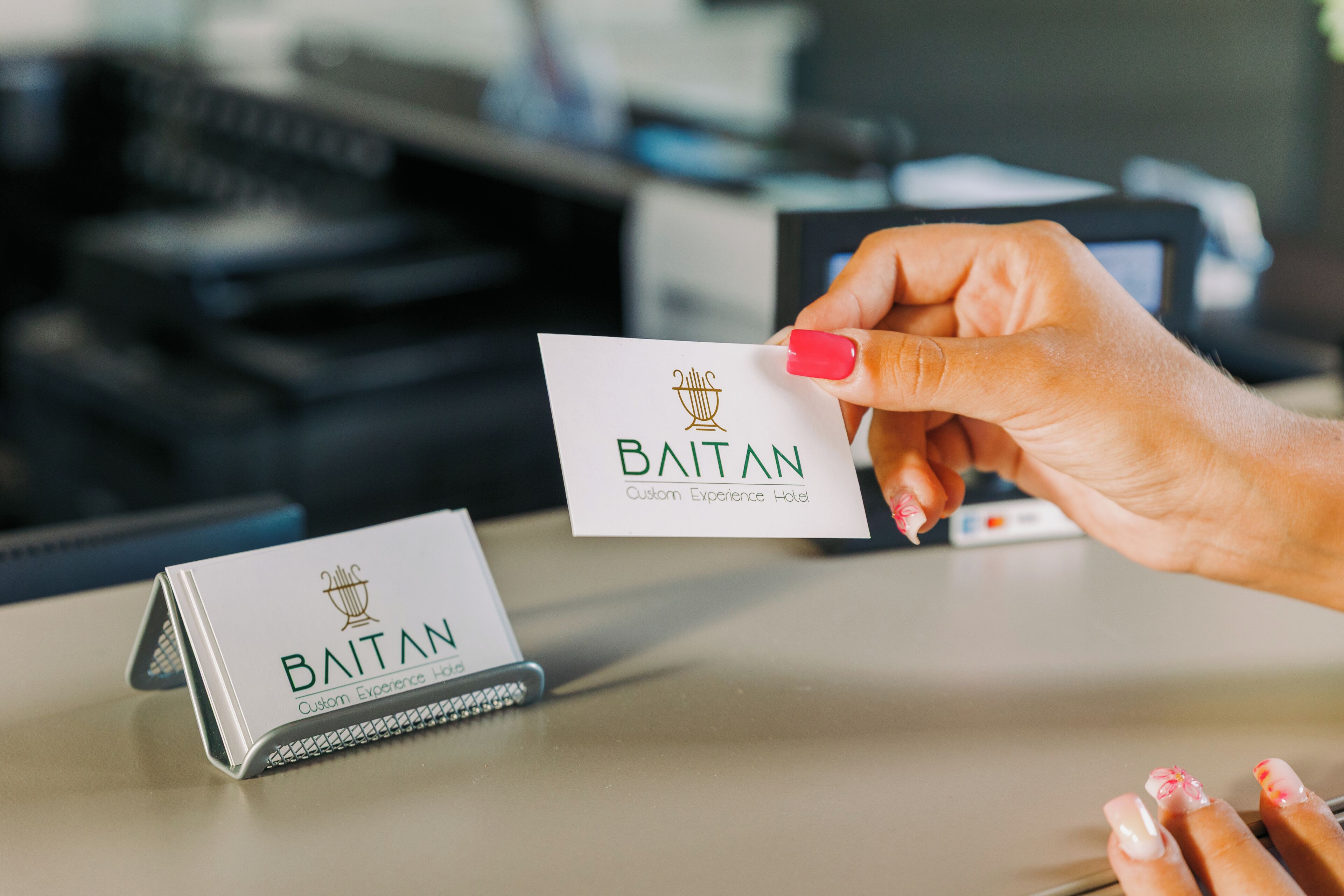 Una mano sostiene una tarjeta de presentación con el nombre 'BAITAN' y el lema 'Custom Experience Hotel' sobre un escritorio.