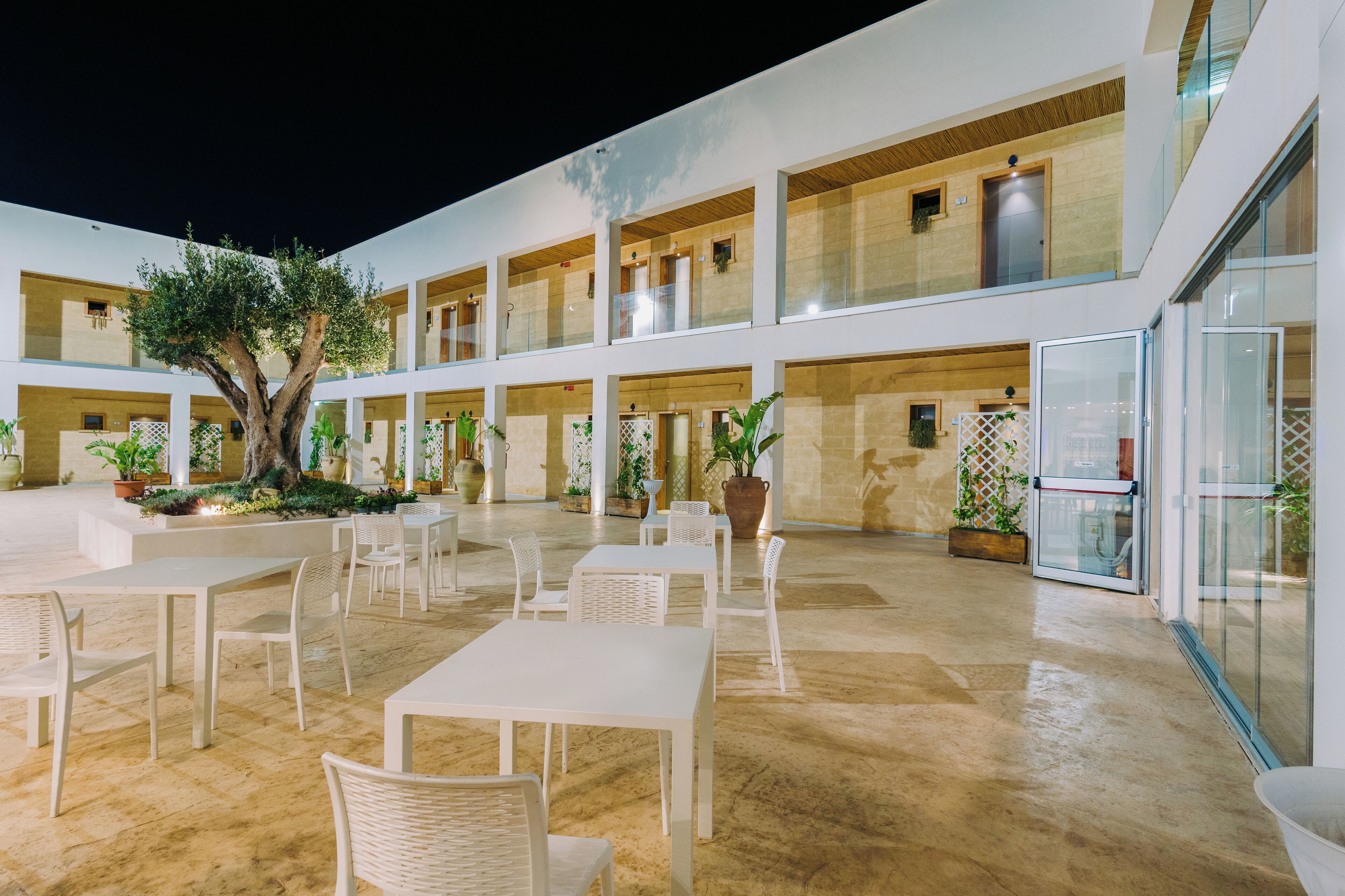 A spacious outdoor area with multiple white tables and chairs, potted plants, and a large tree.