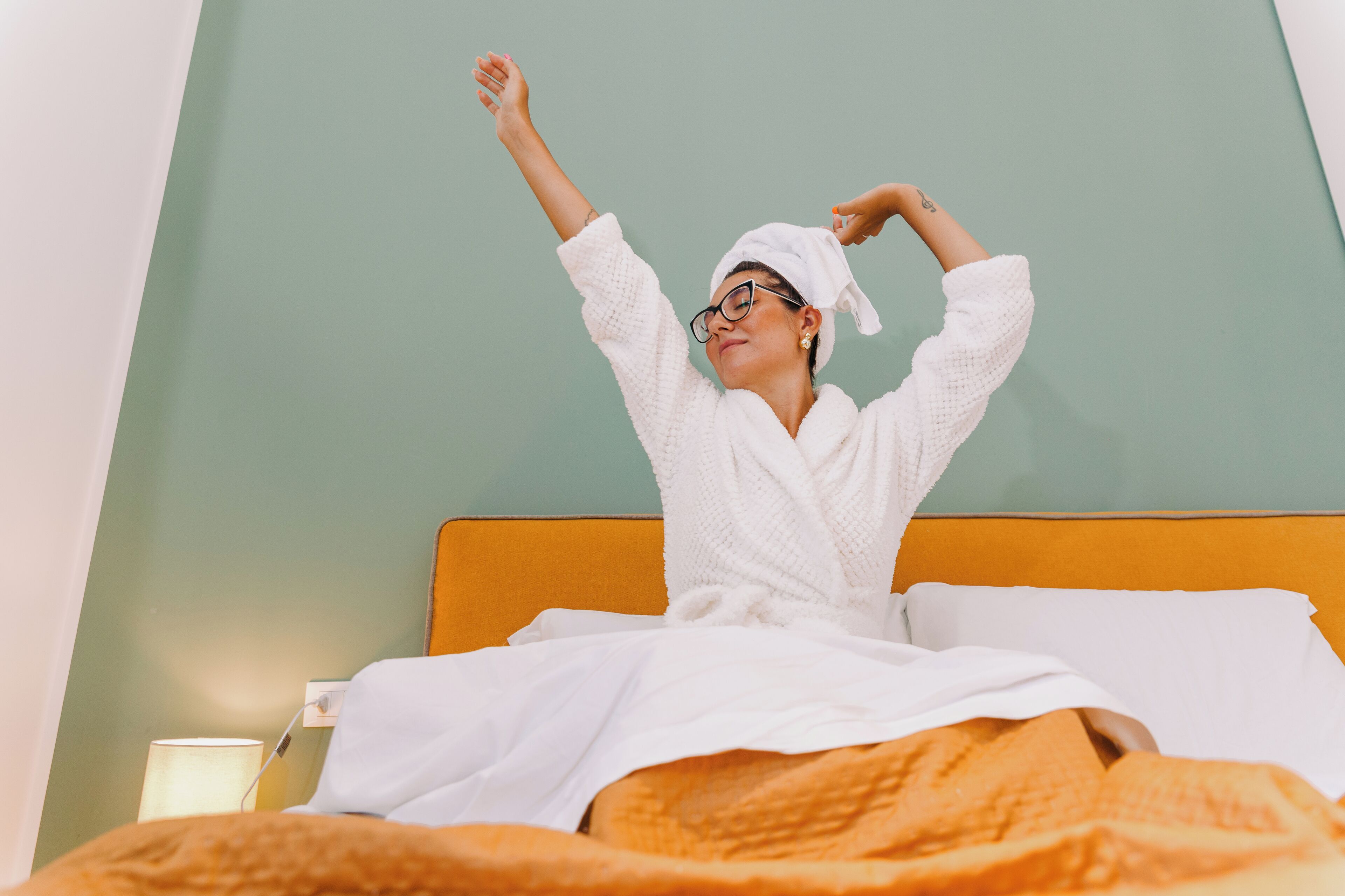 A person in a white bathrobe sitting on a bed with an orange headboard, stretching their arms upwards.