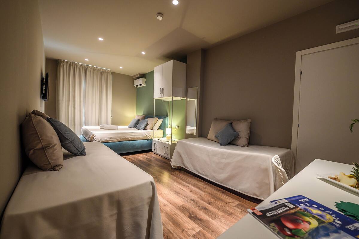 A hotel room with two single beds, a small table with a magazine, a TV mounted on the wall, and a window with curtains.