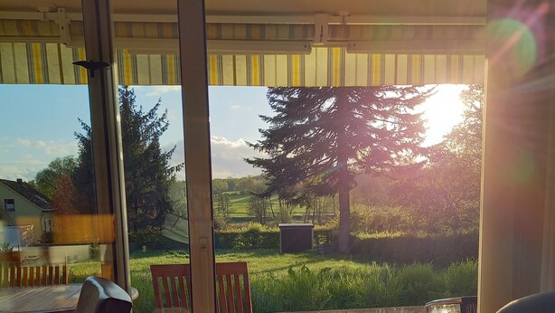 A room with a sliding glass door leading to an outdoor patio with a table and chairs, a view of a tree, and a grassy area. - Erholen, Entspannen, Genießen und Entdecken Eine Oase der Ruhe im Westerwald (Goddert)