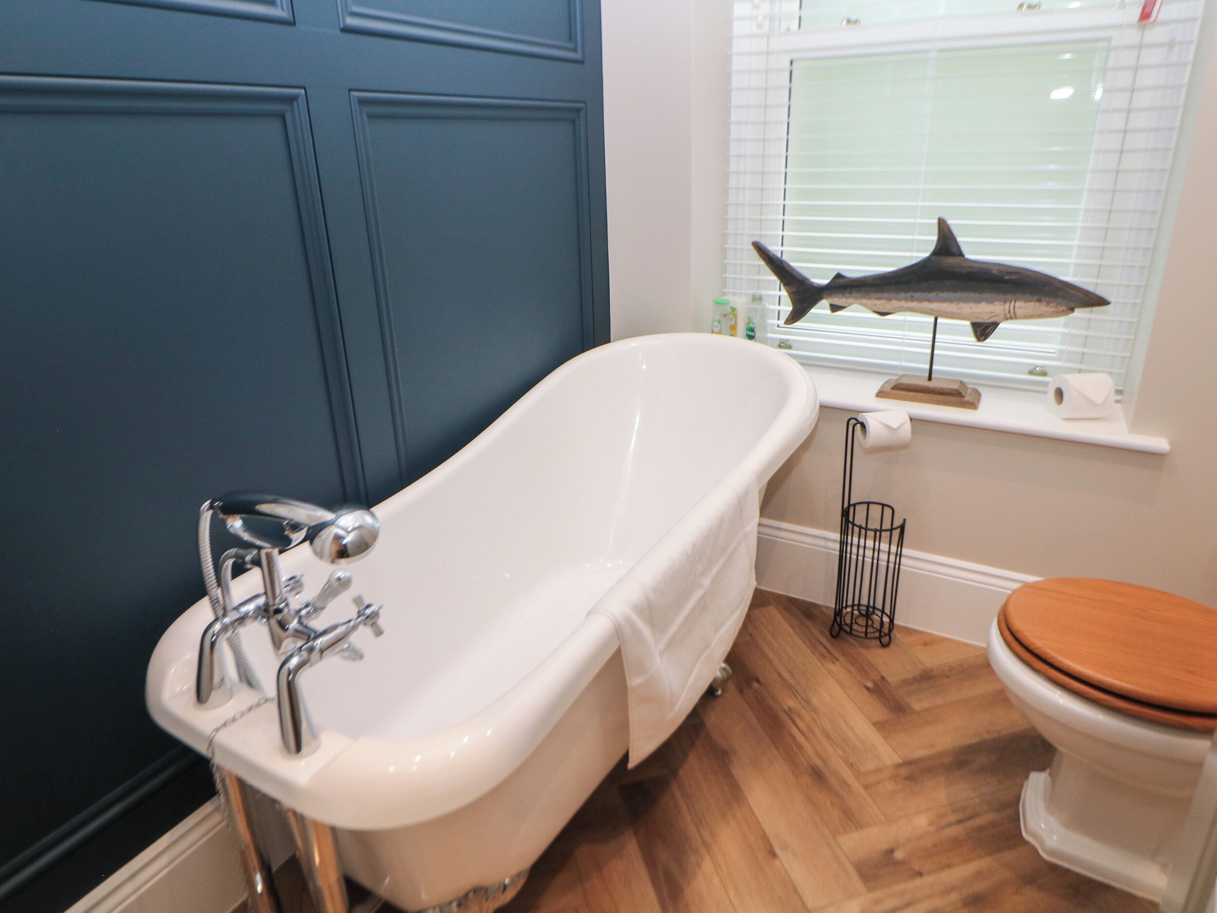 A bathroom with a white bathtub, wooden flooring, a toilet, and a window with blinds.