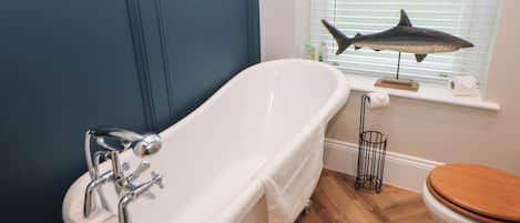 A bathroom with a white bathtub, wooden flooring, a toilet, and a window with blinds.
