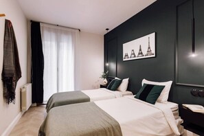 A hotel room with two beds, a large window with sheer curtains, a wall-mounted artwork of the Eiffel Tower, and a bedside table with a vase of flowers.