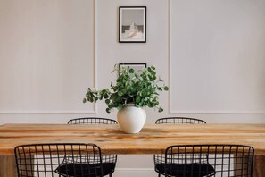 A dining table with a white vase of green plants, black wire chairs, and a framed picture on the wall.