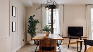 A modern dining area with a wooden table, black wire chairs, a large potted plant, and a TV on the wall. - Lunalux - Habitat Apartments (Barcelona)
