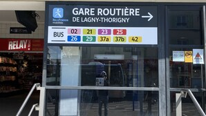 Un panneau d’arrêt de bus indiquant les numéros des lignes, avec un magasin « Relay » en arrière-plan.