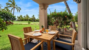 A wooden table set for four with chairs, a tropical garden view, and a clear sky. - Colony Villas Waikoloa 301 (Waikoloa)