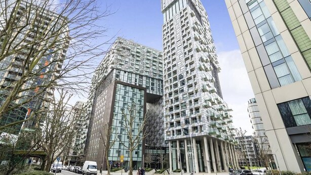A modern high-rise building with a unique facade, surrounded by other tall buildings and trees. - Studio Apartment w/ Balcony In Canary Wharf (London)