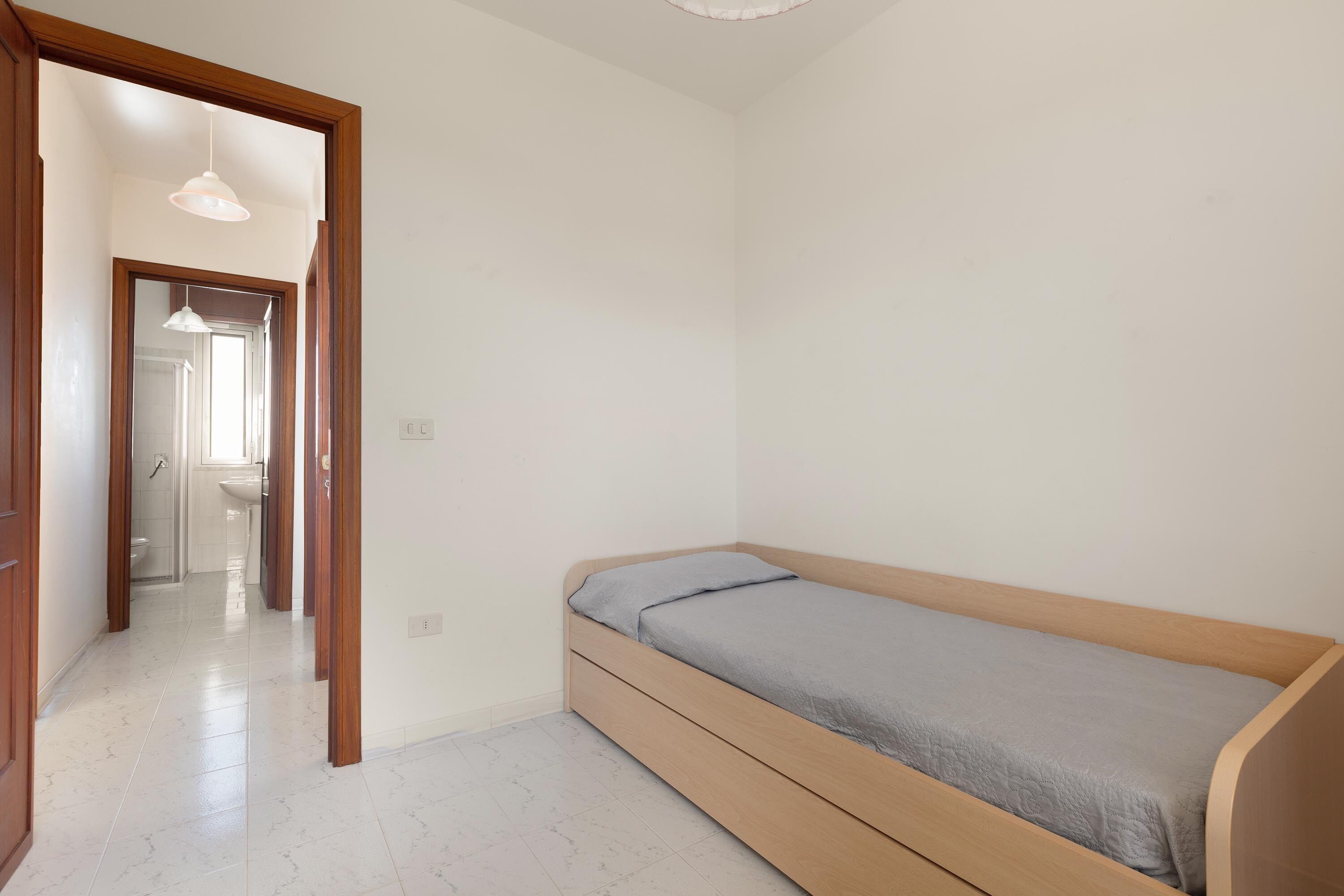 Une chambre avec un lit simple, une tête de lit en bois et un sol carrelé de carreaux blancs.