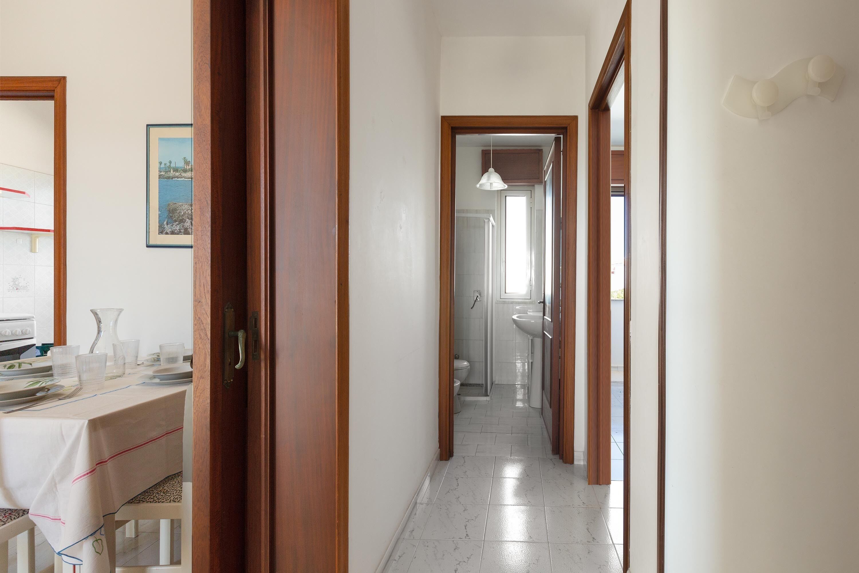 A hallway leading to a bathroom with a toilet, sink, and shower. There is a dining area with a table and chairs visible in the background.