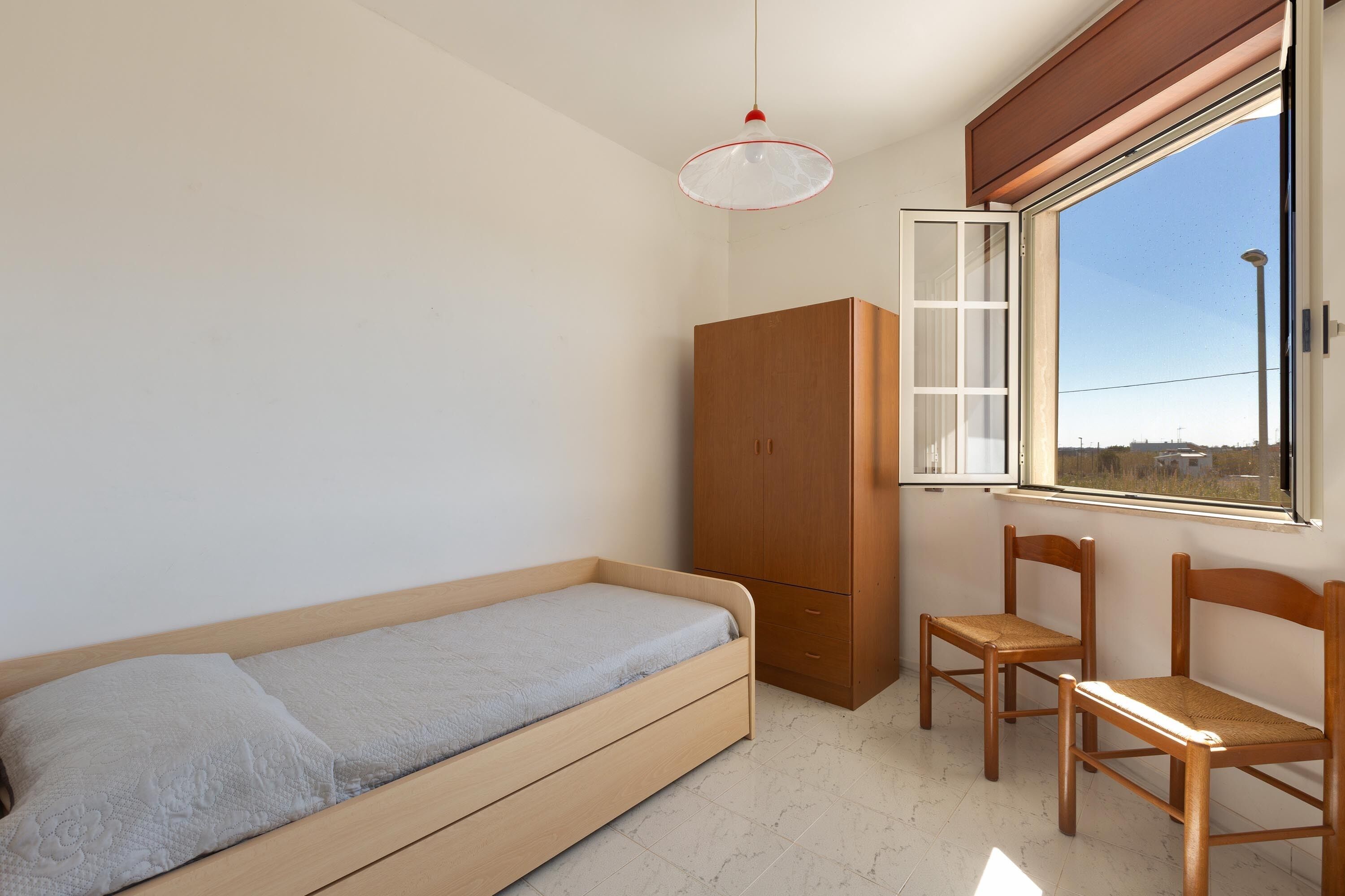 A small, bright room with a single bed, a wooden wardrobe, two chairs, and a window offering a view of the outside.
