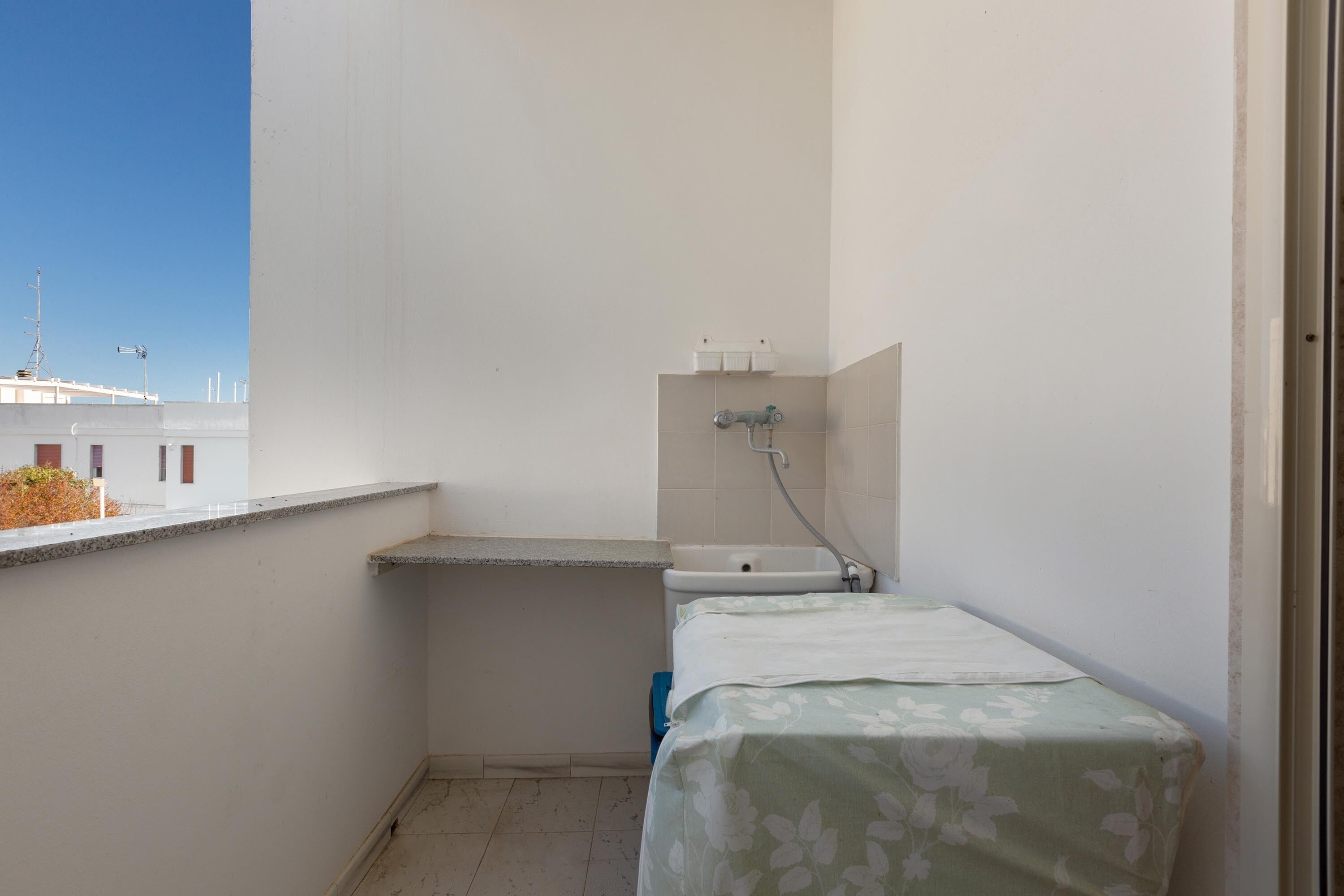 A small, narrow balcony with a single bed, a sink, and a window overlooking a building.