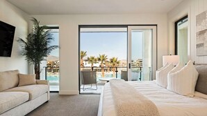 Camera d'hotel moderna con un letto grande, una poltrona, una pianta in vaso e vista su piscina e palme.