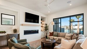Un soggiorno moderno con divano, poltrone, tavolino e TV a parete. Si vede un'ampia finestra con vista su palme e edifici.