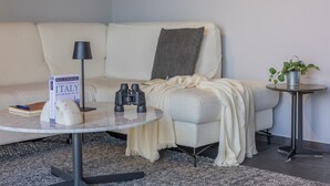 Una sala de estar moderna con un sofá blanco, una mesa redonda con un libro y unos binoculares, una lámpara y una planta pequeña sobre una mesita auxiliar.