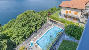 Una piscina con vistas a un lago y montañas, un edificio con tejado de tejas rojas y un balcón con mobiliario exterior.
