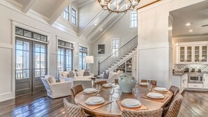 A spacious living room with a high ceiling, large windows, and a dining area set for six.