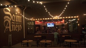 A bar area with a large screen displaying a sports event, string lights, and several tables and chairs.