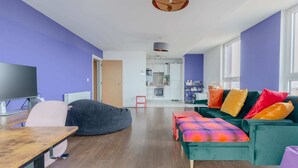 Ein modernes Wohnzimmer mit einem großen Fernseher, einem Sitzsack, einer Couch mit bunten Kissen und einer Küche im Hintergrund.