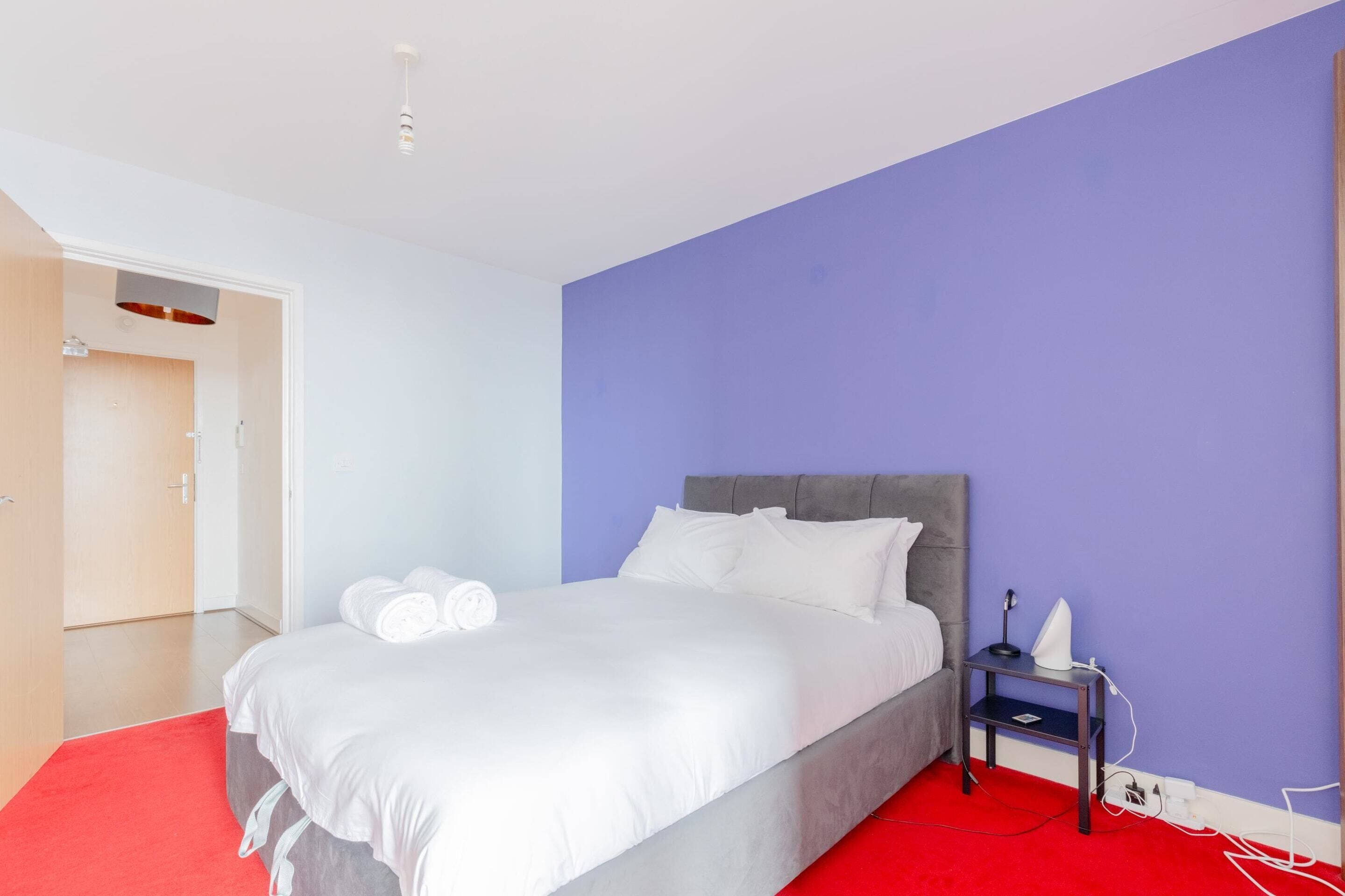 Quarto moderno com cama cinza, roupa de cama branca, tapete vermelho, mesa de cabeceira com abajur e parede azul.