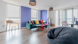 Ein modernes Wohnzimmer mit einem grünen Sofa, einem roten Couchtisch und einem großen Sitzsack. Große Fenster sorgen für natürliches Licht und bieten Blick auf die Stadt.