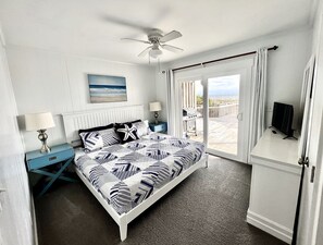 A bedroom with a bed, a ceiling fan, a nightstand, a TV, and a view of the beach through a sliding door.