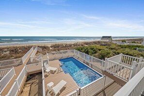 A swimming pool with a wooden deck, white railings, and a view of the beach.