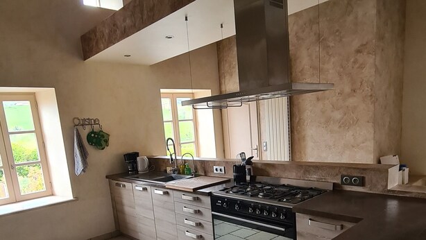 A spacious kitchen with a large island, a built-in oven, and a stainless steel range hood. There is a window providing natural light and a view of greenery outside.