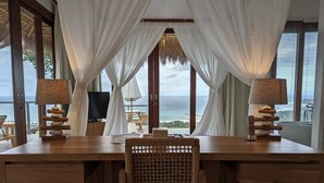 Uma mesa de madeira com uma cadeira de fibra natural, duas luminárias, uma televisão e vista para o oceano através de cortinas leves.
