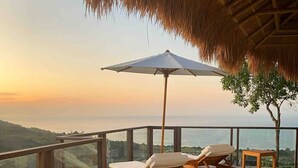 Um deck de madeira com espreguiçadeiras e uma mesa, com vista para um corpo d'água e colinas.