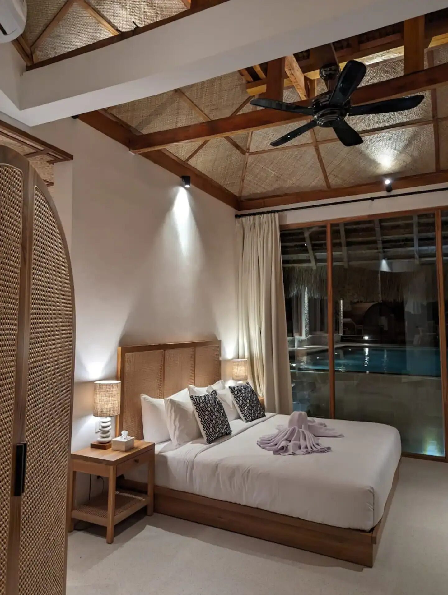 Une chambre dotée d’un grand lit, d’une table de chevet, d’un ventilateur de plafond et offrant une vue sur une piscine extérieure.