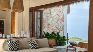 Uma sala de estar moderna com sofá, mesa de centro de madeira e cadeiras. Há uma parede de pedra e vista para o oceano.