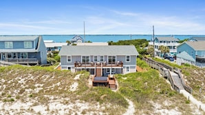 Une propriété située en bord de mer, avec une grande maison dotée d’une terrasse et de nombreuses fenêtres, entourée de dunes de sable et offrant une vue sur l’océan.