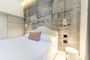 Een slaapkamer met een bed, witte beddengoed, een gestreept kussen, een wandplank en een witte nachtkastje met een lamp.