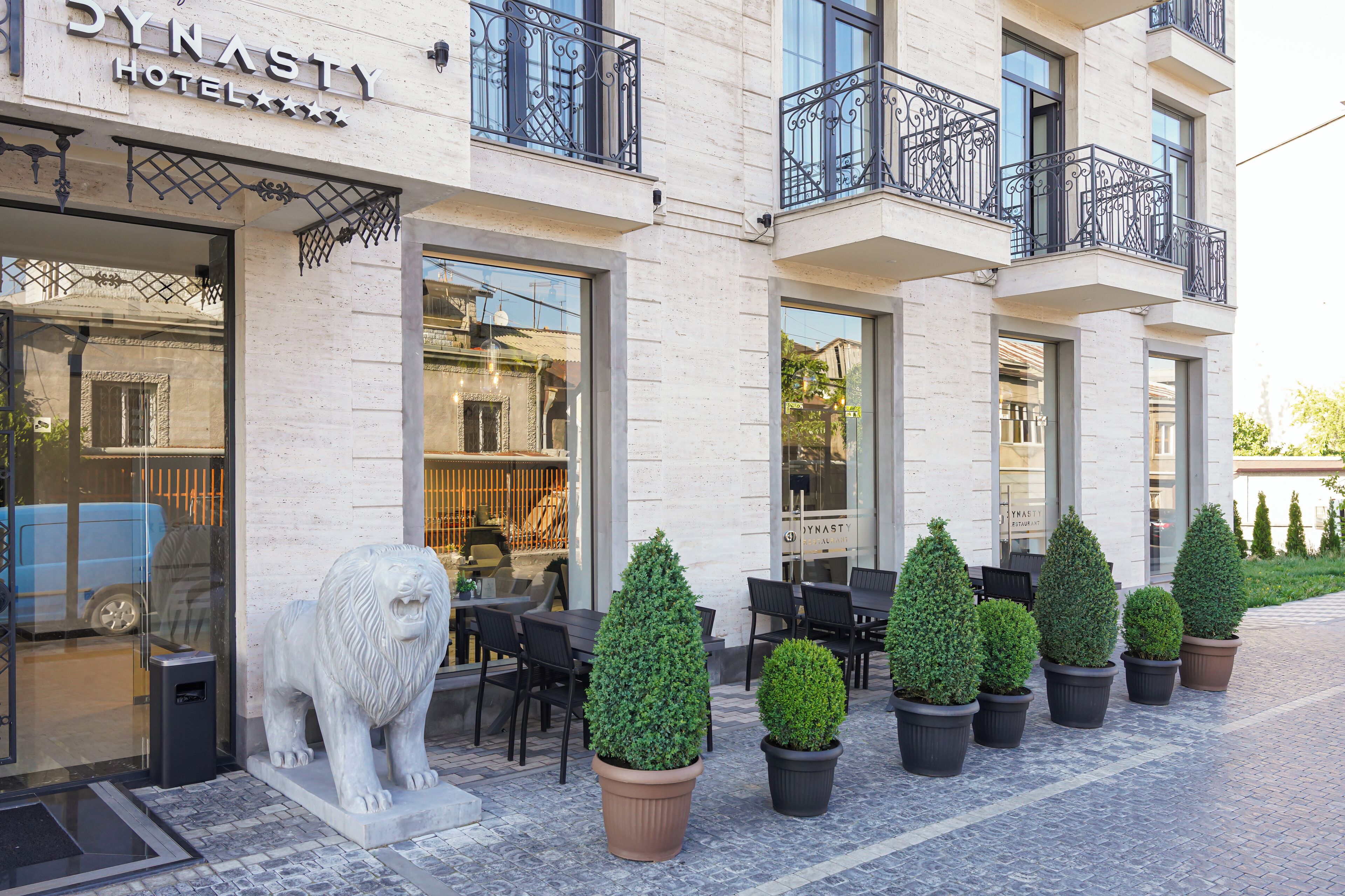 A hotel exterior with a stone facade, outdoor seating area with tables and chairs, and a lion statue in front.