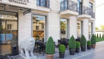 A hotel exterior with a stone facade, outdoor seating area with tables and chairs, and a lion statue in front.