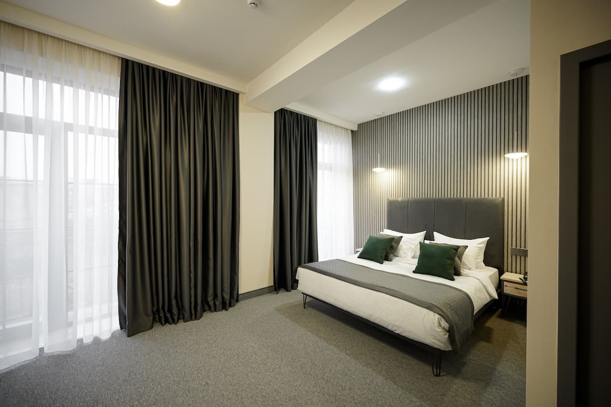 A modern hotel room with a large bed, dark curtains, a headboard with vertical lines, and a bedside table with a lamp.